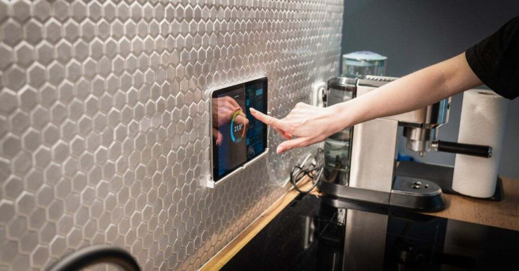 Person using a smart home control panel in a modern kitchen to manage lighting, temperature, and connected devices.